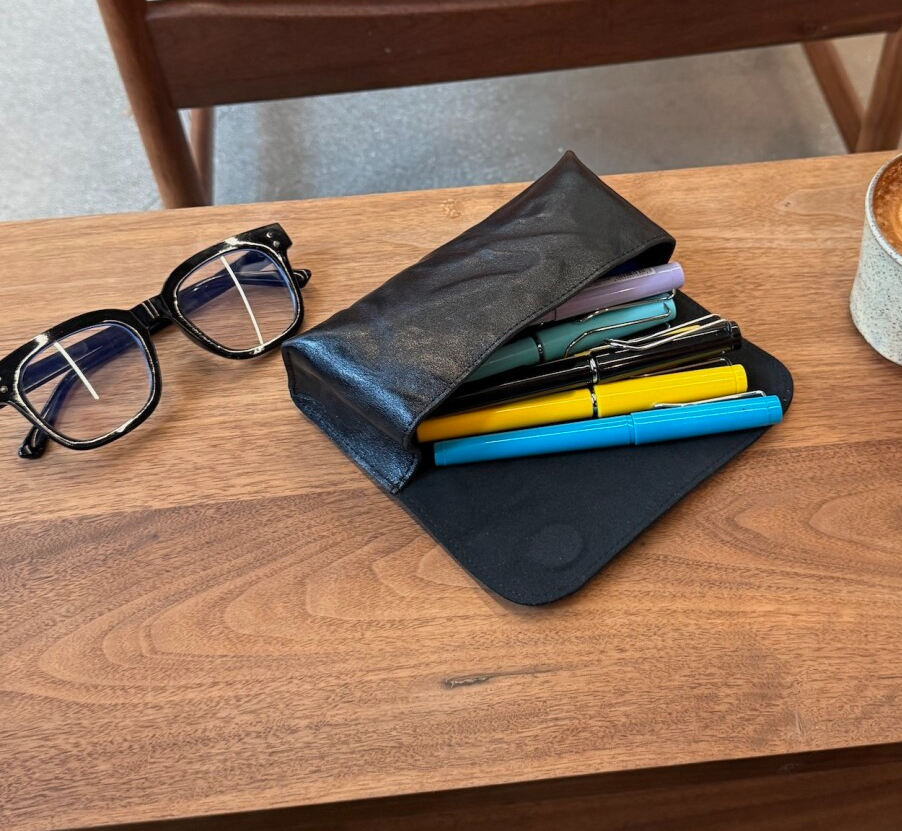 Black pouch with stationery items on a wooden table with a cup of coffee and glasses.