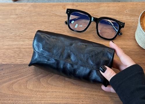 Black glasses case and black glasses on a wooden table with a coffee cup in the background.