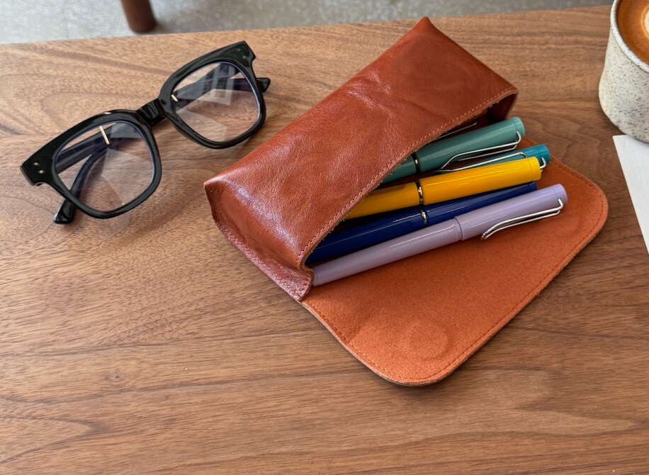 Brown leather case with pens, glasses, and a coffee cup on a wooden table.