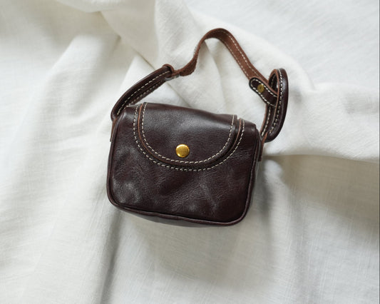 A small dark brown leather shoulder bag with a gold snap button and white contrast stitching, resting on a draped white fabric background.