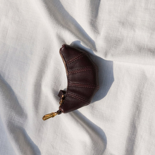 A small dark brown leather pouch with a gold-tone zipper pull, placed on a textured white fabric background.