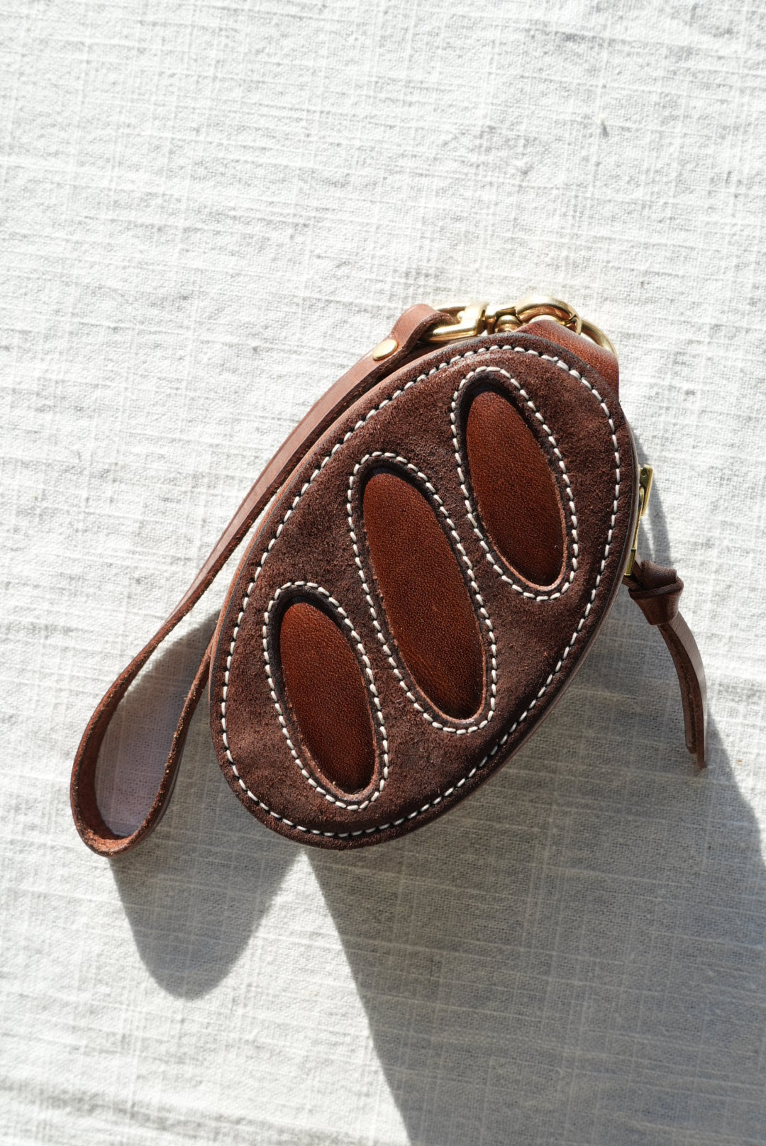 A small brown leather wristlet or coin purse with three oval cutouts on the front, white contrast stitching, and a wrist strap, photographed on a white textured fabric background.