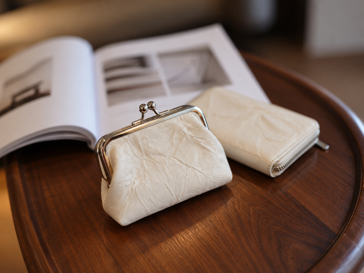 Two cream-colored textured leather coin purses with metal kiss-lock and zipper closures, displayed on a dark wooden table next to an open magazine.